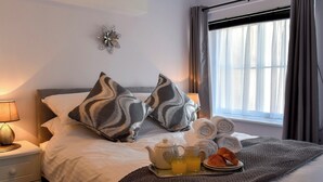 Ein ordentlich bezogenes Bett mit zwei gemusterten Kissen, ein Nachttisch mit Lampe und ein Tablett mit Frühstücksangeboten, darunter ein Teekessel, Tassen und Gebäck.