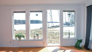 Una habitación con ventanas amplias que dan a un lago, suelo de madera y algunas plantas en macetas.