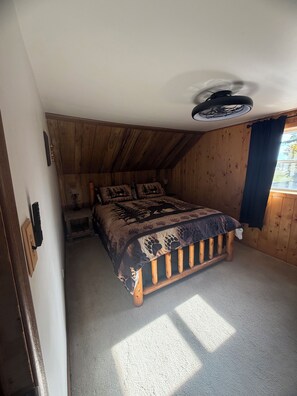 Une chambre avec un plafond en pente, des murs en bois, un lit recouvert d’une couvre-lit à motifs, une petite table de chevet et un ventilateur de plafond.