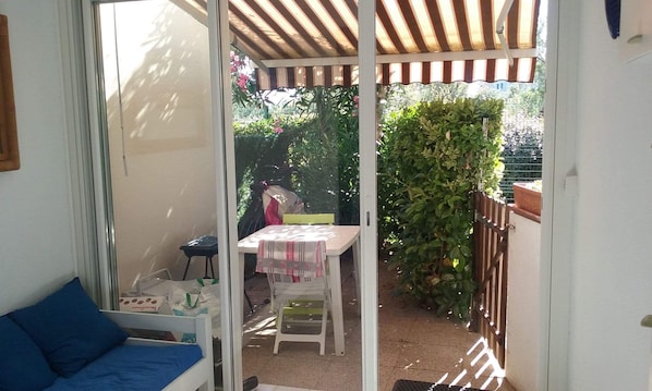 A room with a bed, a small table with a tablecloth, and a chair. There is a window with a view of a garden outside. - Apartment Cap D'Agde, Mail Rochelongue (Cap d agde)