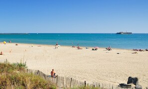 A wide sandy beach with a wooden fence in the foreground, a clear blue sky, and calm sea.