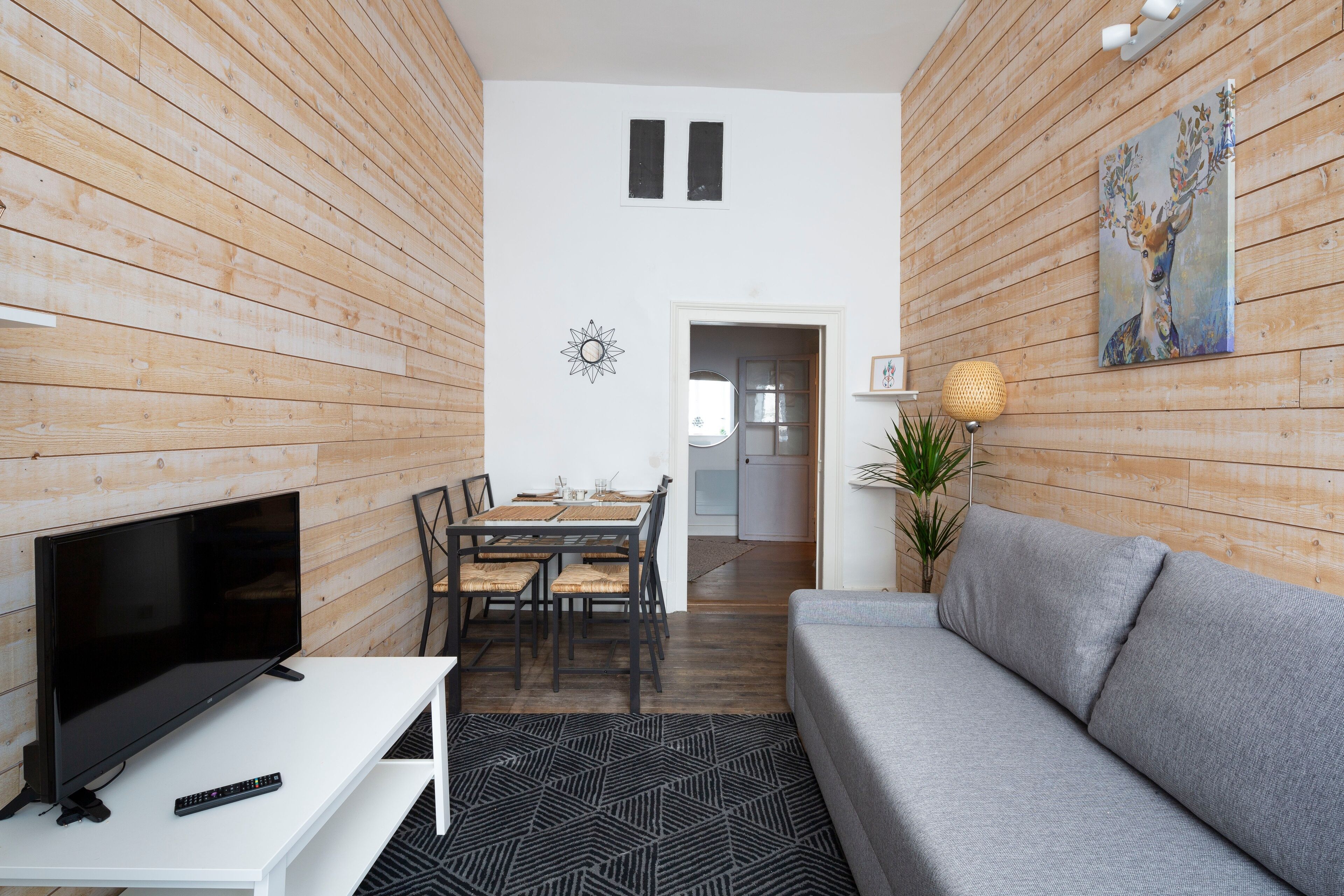 A modern living room with a flat-screen TV, a grey sofa, and a dining area with a wooden table and chairs.