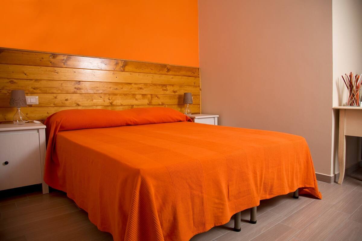 A room with a wooden headboard, an orange bedspread, two bedside tables with lamps, and a small table with decorative items.