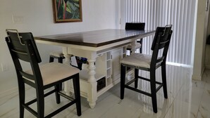 Un comedor con una mesa de madera y sillas negras, un cuadro de loros en la pared, y una ventana con persianas.