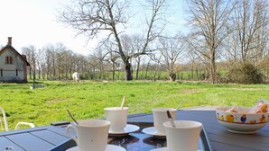 Ein Tablett mit vier weißen Tassen und Untertassen auf einem Tisch, eine Schüssel mit Brot und im Hintergrund eine Wiese mit Bäumen und einem Haus.