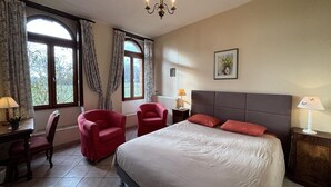 Ein Schlafzimmer mit einem großen Bett, zwei roten Sesseln, einem Holztisch, einer Kandelaber und großen Fenstern mit Vorhängen.