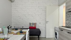 Un espace de vie compact avec un mur en briques blanches, un lit agrémenté d’un coussin coloré, une table à manger avec un set de table coloré, un lave-linge et une petite kitchenette.