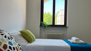 Une chambre avec deux lits, une fenêtre donnant sur des immeubles et des arbres, un radiateur et une serviette sur le lit.