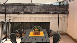 Une terrasse extérieure avec une table et des chaises, un panier de fleurs et un mur en béton en arrière-plan.