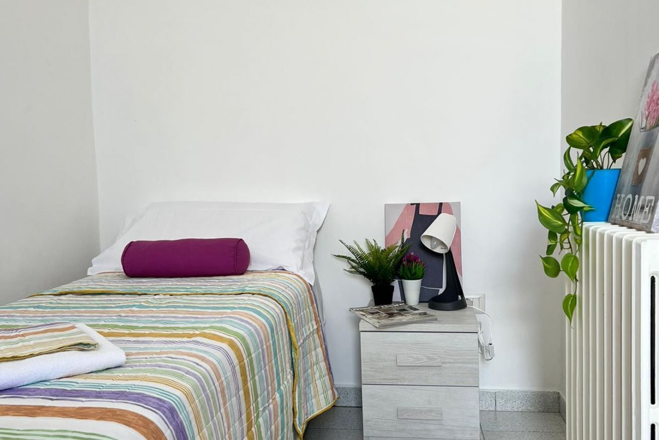 Un dormitorio ordenado con cama, mesita de noche con lámpara, pequeña planta en maceta y radiador.