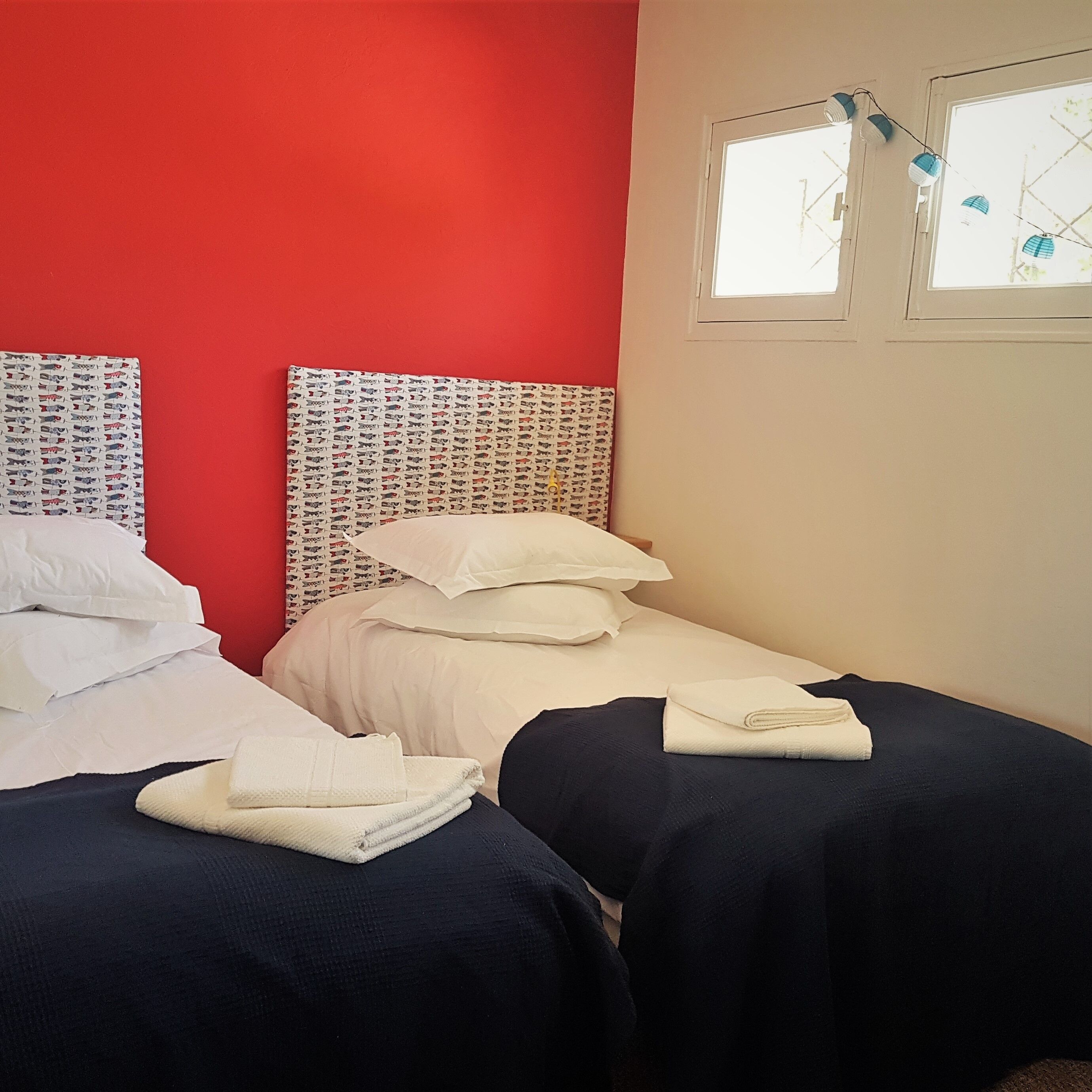 Habitación con dos camas, cada una con ropa blanca y sábanas azul oscuro. Las paredes son rojas y hay una ventana con vistas a árboles.