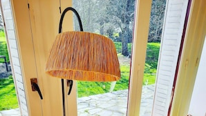 Una habitación con un sofá verde, una pequeña mesa de madera con un libro, una lámpara de pie y vistas a un sendero de piedra y vegetación al exterior.