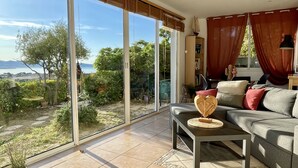 Un salon spacieux doté d’une grande porte-fenêtre coulissante donnant sur un jardin, d’un canapé gris avec des coussins et d’une petite table en bois.