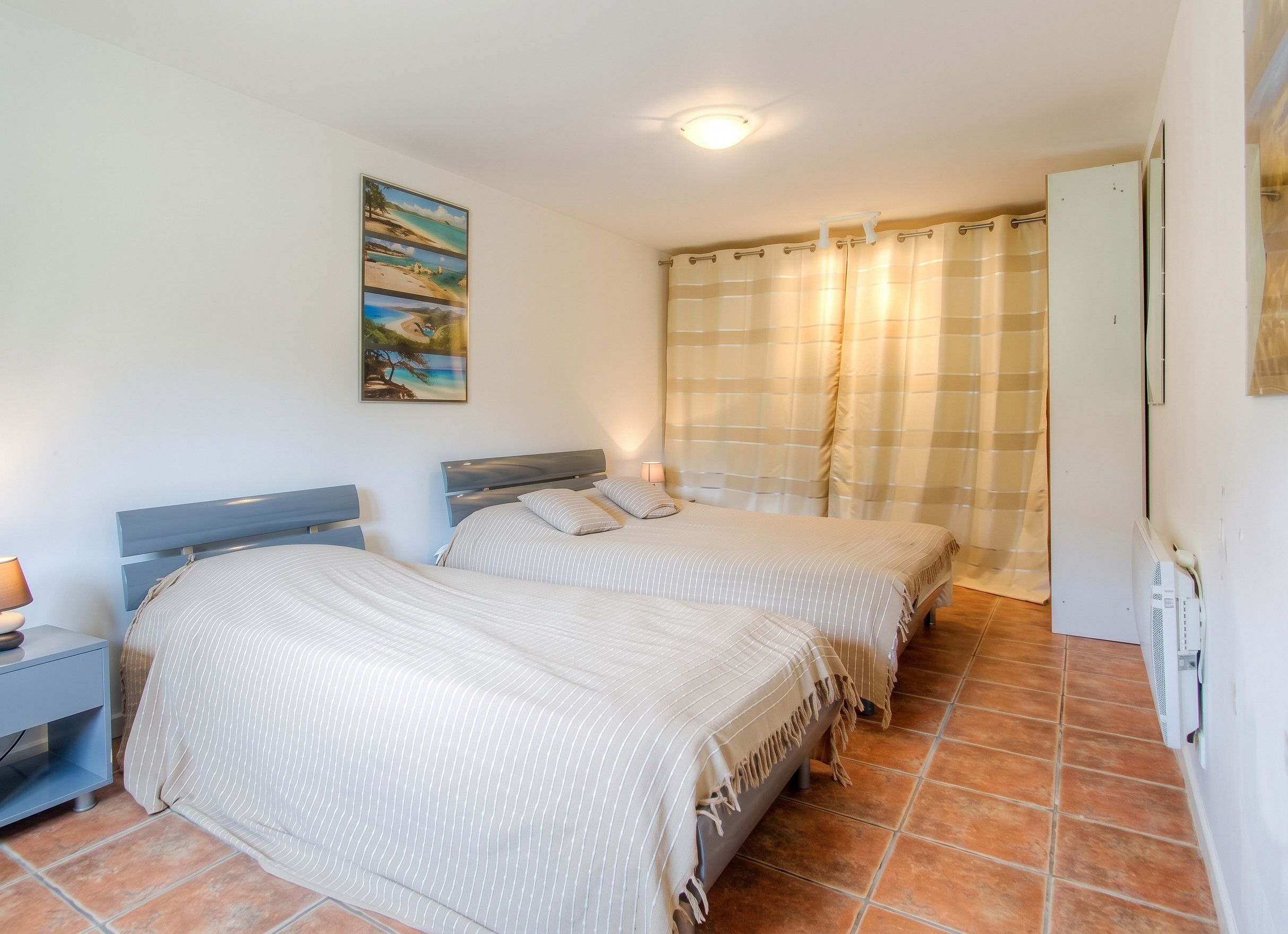 A bedroom with two beds, a nightstand, a lamp, a painting, and a bathroom with a shower curtain.