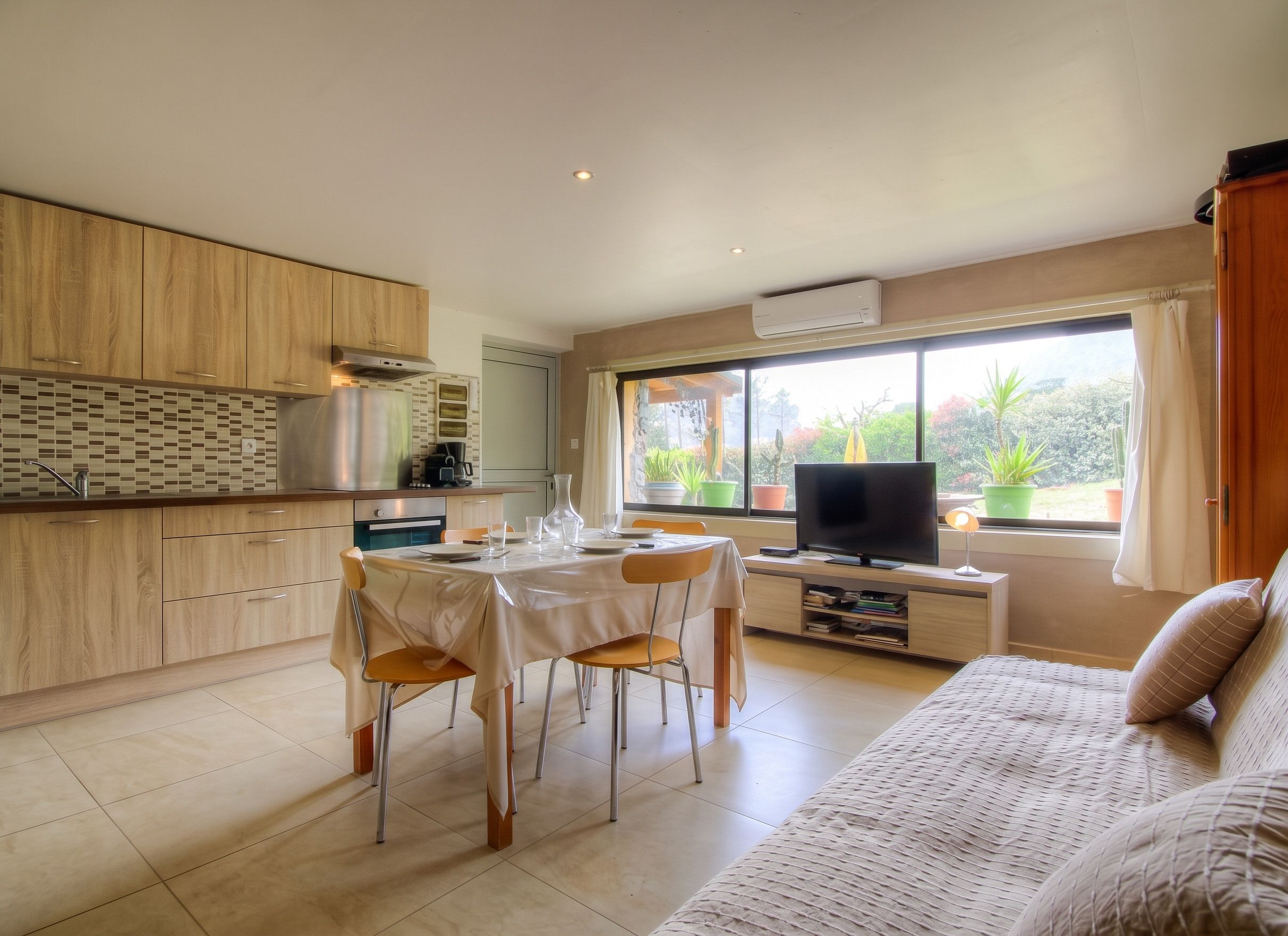 A modern kitchen with wooden cabinets, a dining area with a table and chairs, a TV, and a large window with a view of greenery.