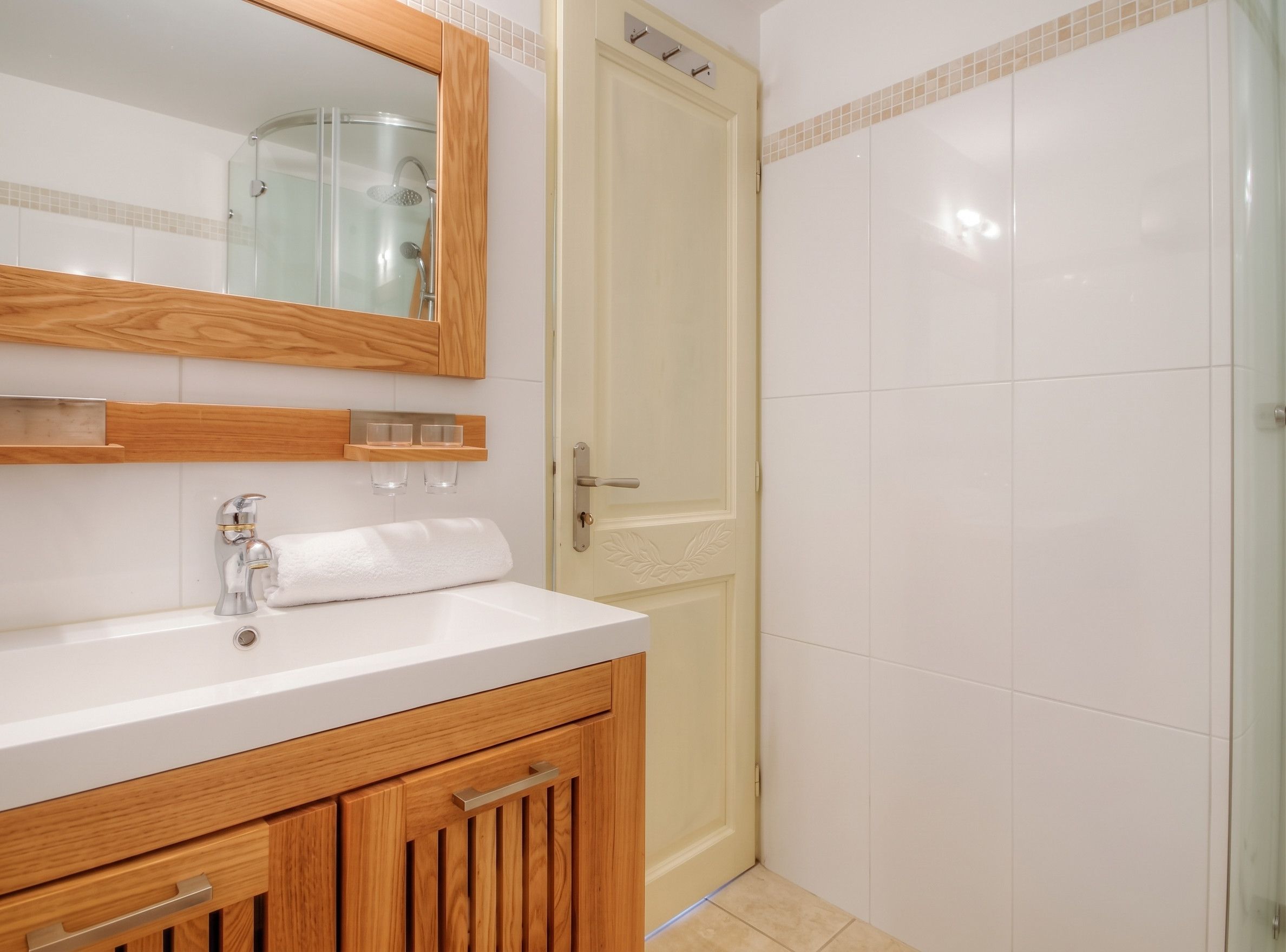 Une salle de bain avec un lavabo blanc, un meuble-lavabo en bois, une cabine de douche en verre et une porte dotée d’une poignée.