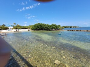 A beach with clear shallow waters, a thatched-roof hut, and a white building with an American flag.