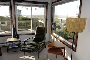 Quarto com vista para a praia, uma cadeira verde, uma mesa pequena e um abajur.