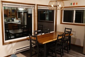 Un comedor con una mesa y sillas de madera, ventanas amplias y una cocina visible a través del vidrio.