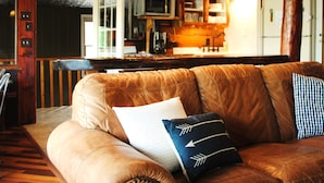 Un salon chaleureux avec un canapé en cuir marron, deux coussins à motifs et un plafond en bois avec des poutres apparentes.