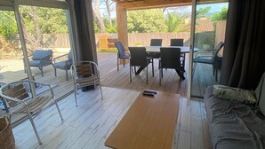 Amplio espacio interior con piso de madera, un sofá, una mesa y sillas. Hay una puerta corrediza de vidrio que da a un patio exterior con más asientos.