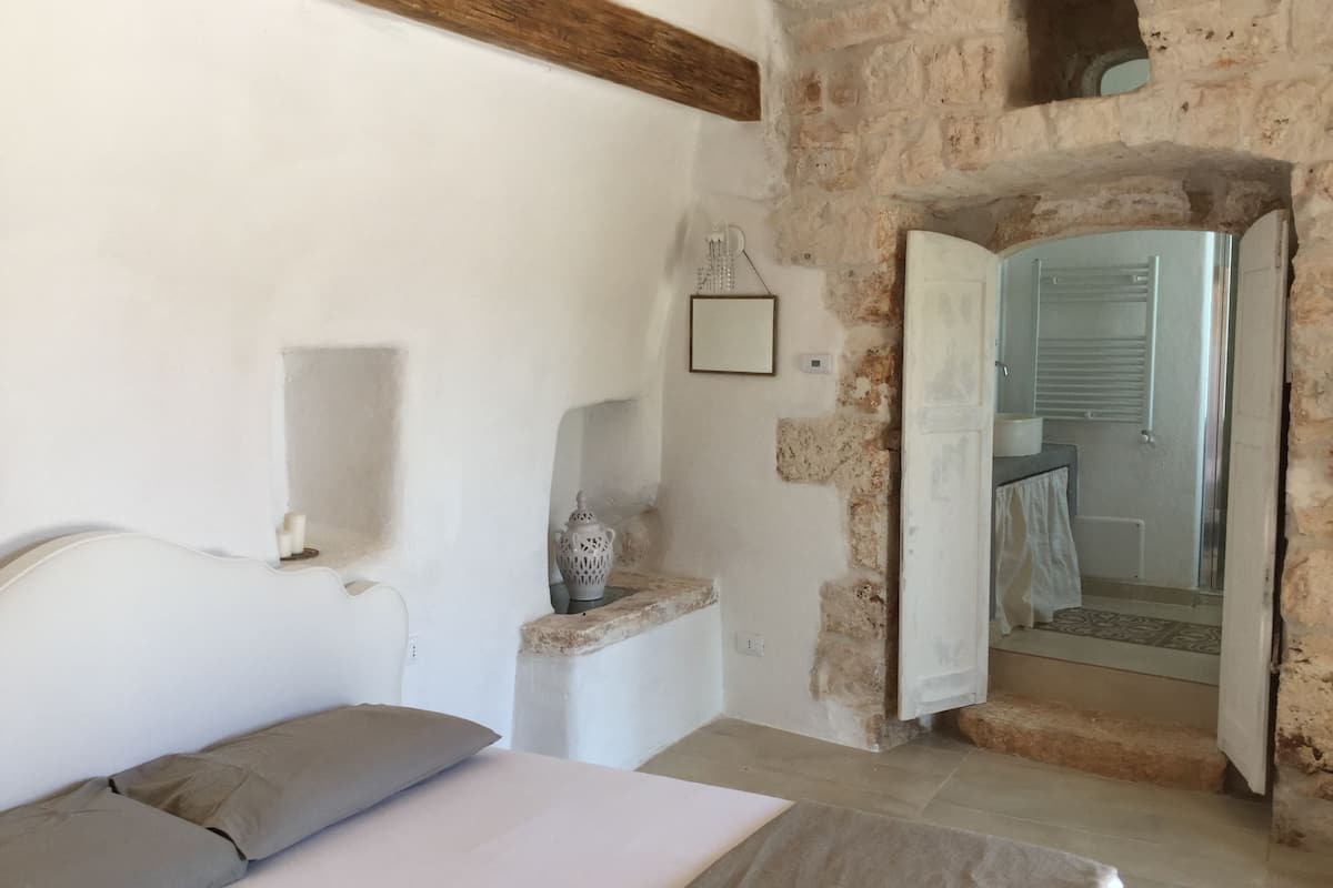 A bedroom with a bed, a nightstand, a stone wall, and a bathroom visible through an open door.