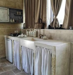 A kitchen with a stone wall, a built-in shelf with kitchenware, and a window with curtains. - Relais Grottone (Ostuni)