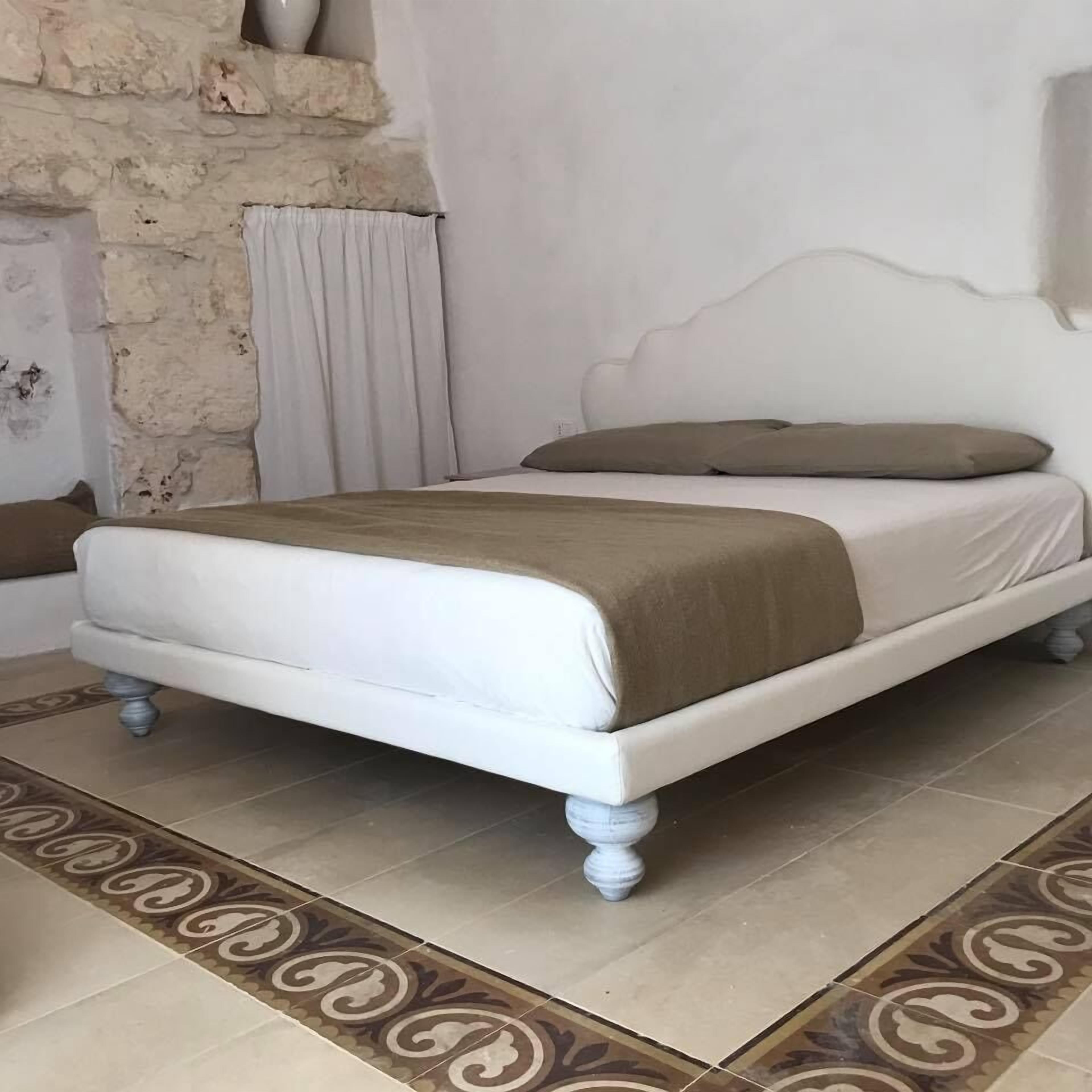 Una cama con cabecera blanca, una colcha color beige y un almohadón marrón en una habitación con baldosas de suelo estampadas y pared de piedra.