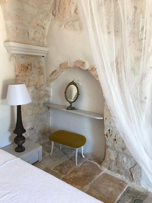 Un dormitorio con pared de piedra, una cama blanca, una mesita de noche con lámpara, un banco y un espejo sobre una estantería.