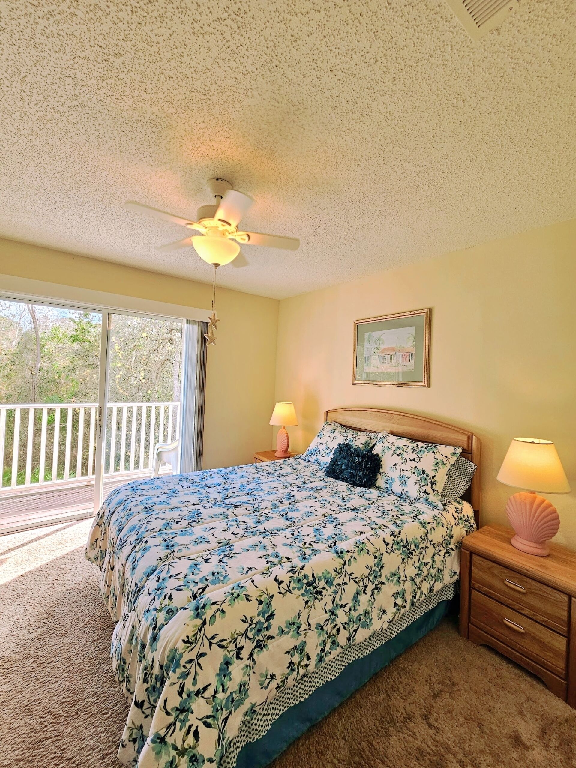A bedroom with a bed, two bedside tables with lamps, a ceiling fan, a window with a view of greenery, and a framed picture on the wall.