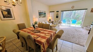 Uma sala de jantar com uma mesa posta para quatro pessoas, rodeada por cadeiras de vime. Há um candelabro sobre a mesa, e uma pintura é visível na parede à esquerda.