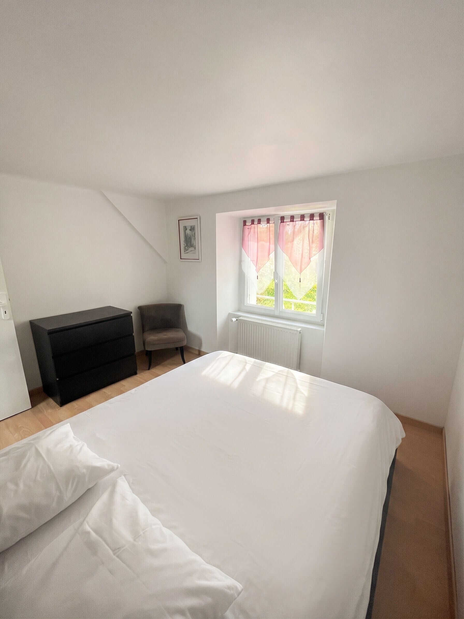 Un dormitorio con cama, cómoda, silla, ventana con cortinas y radiador.