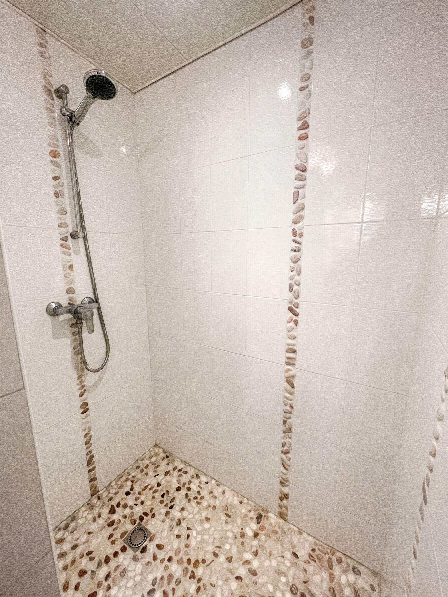 Une douche équipée d’un pommeau de douche à main et d’un robinet mural, d’un sol de douche à motifs de galets et de carreaux décoratifs dans un coin.