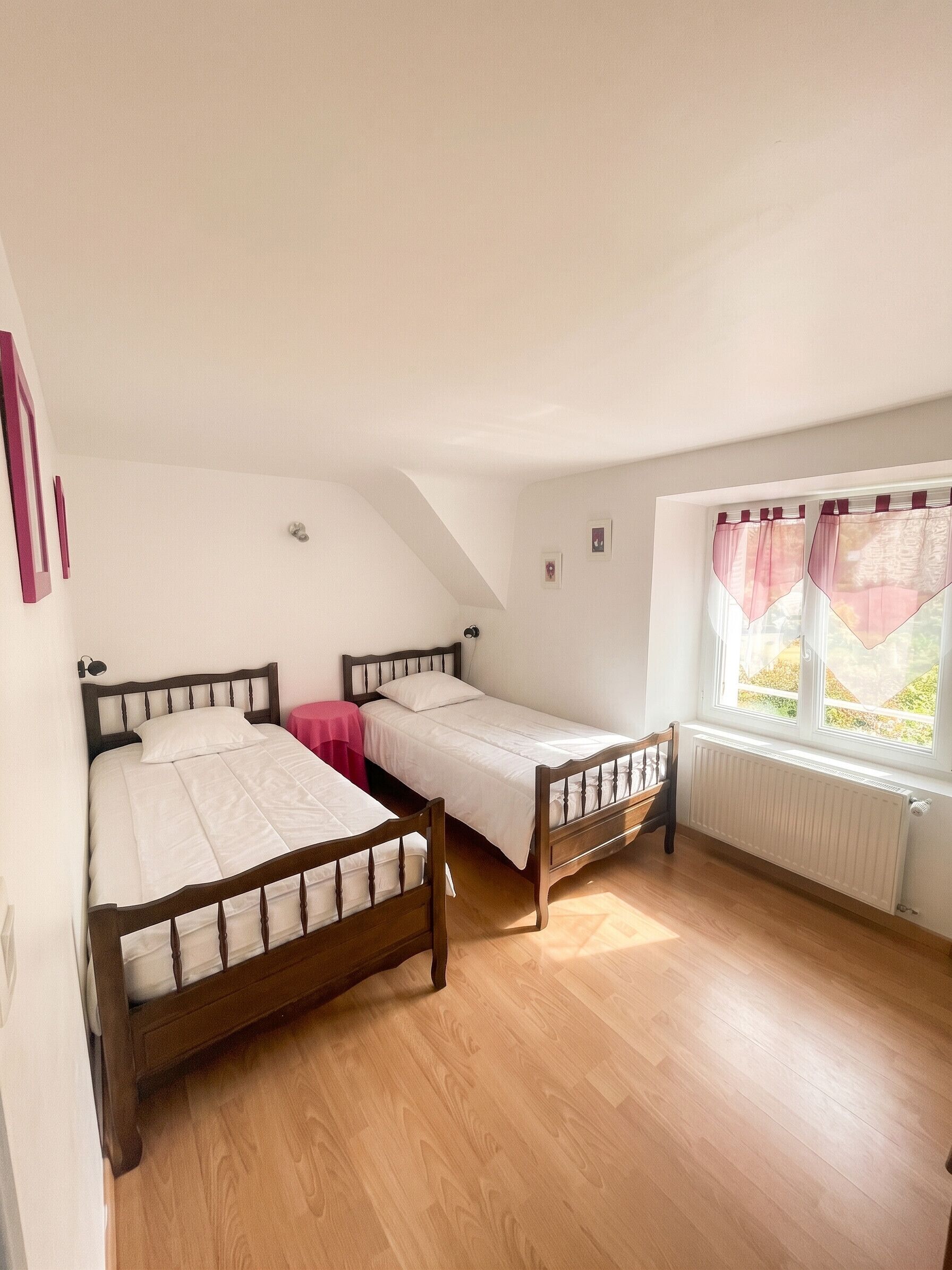 A room with two wooden beds, a window with curtains, a radiator, and a pink table.