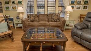 Una sala de estar con mesa de fútbolín, un sofá marrón, un sillón gris y una mesa de centro de madera.