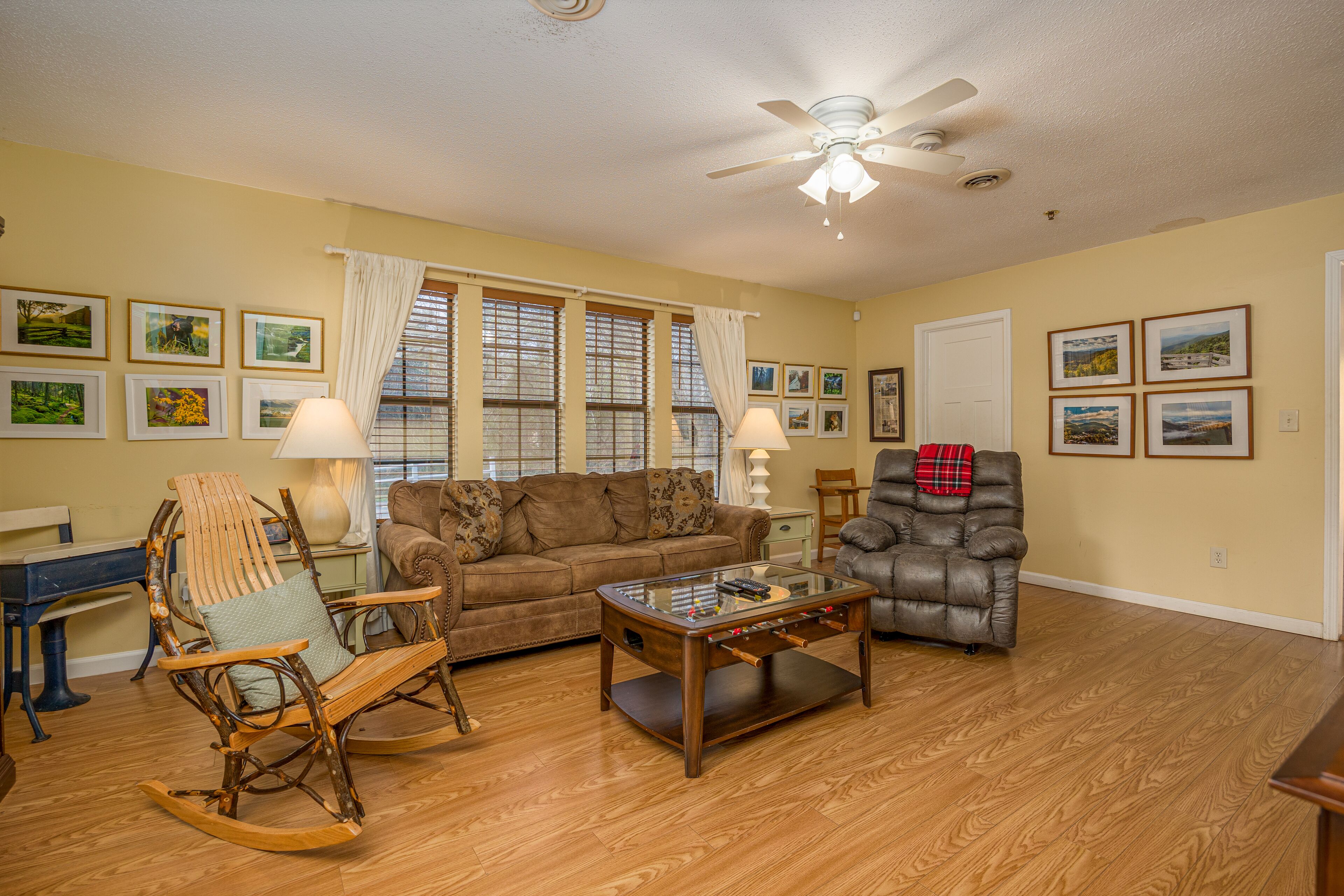 A living room with a wooden floor, a brown sofa, a leather armchair, a glass coffee table, and a ceiling fan.