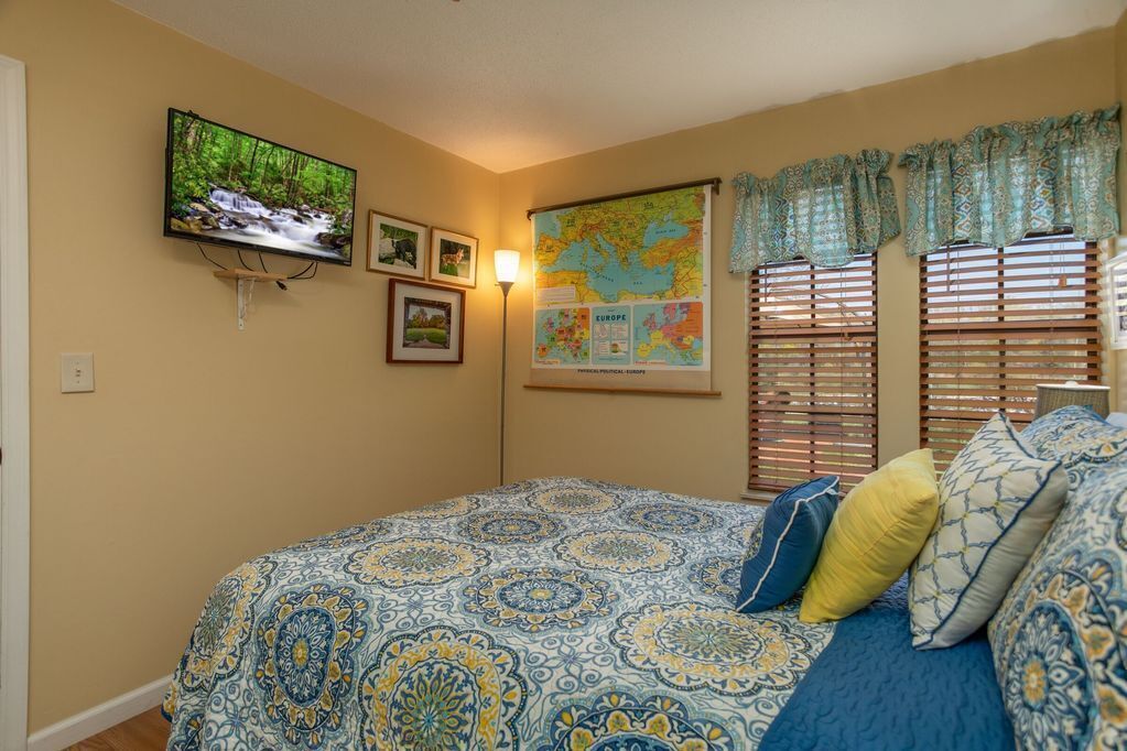 A bedroom with a bed, a television, a map, and a window with blinds.