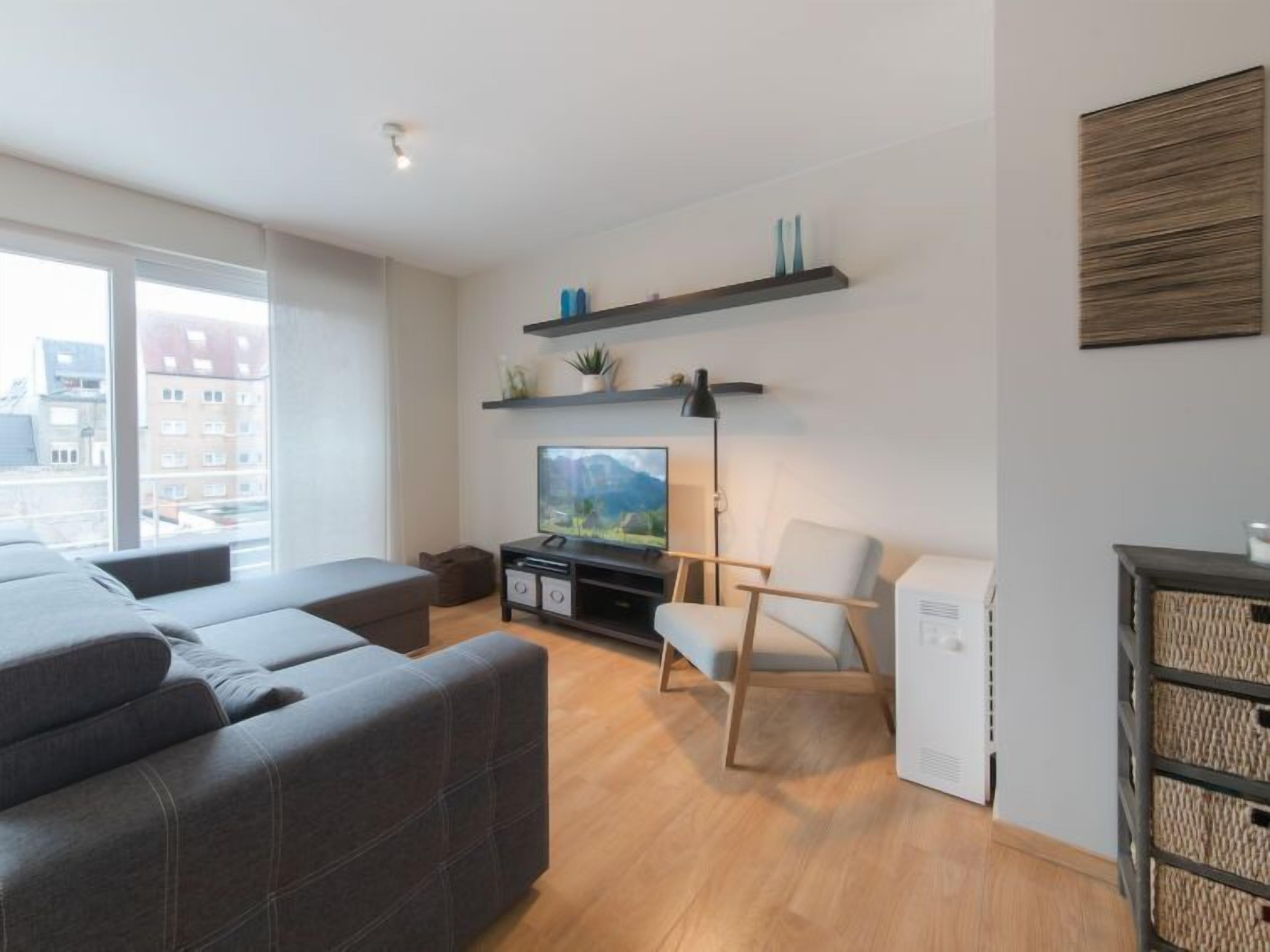 Una sala de estar moderna con un sofá, un televisor, una mesita y una silla. Hay estanterías sobre el televisor y una lámpara en el suelo.