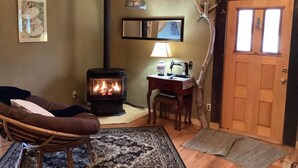 Un salon chaleureux avec un poêle à bois, un fauteuil à bascule, un bureau avec une machine à coudre et une porte en bois.