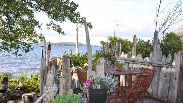 Une terrasse en bois avec une table et des chaises donnant sur une étendue d’eau, entourée de plantes en pot et d’une clôture rustique.