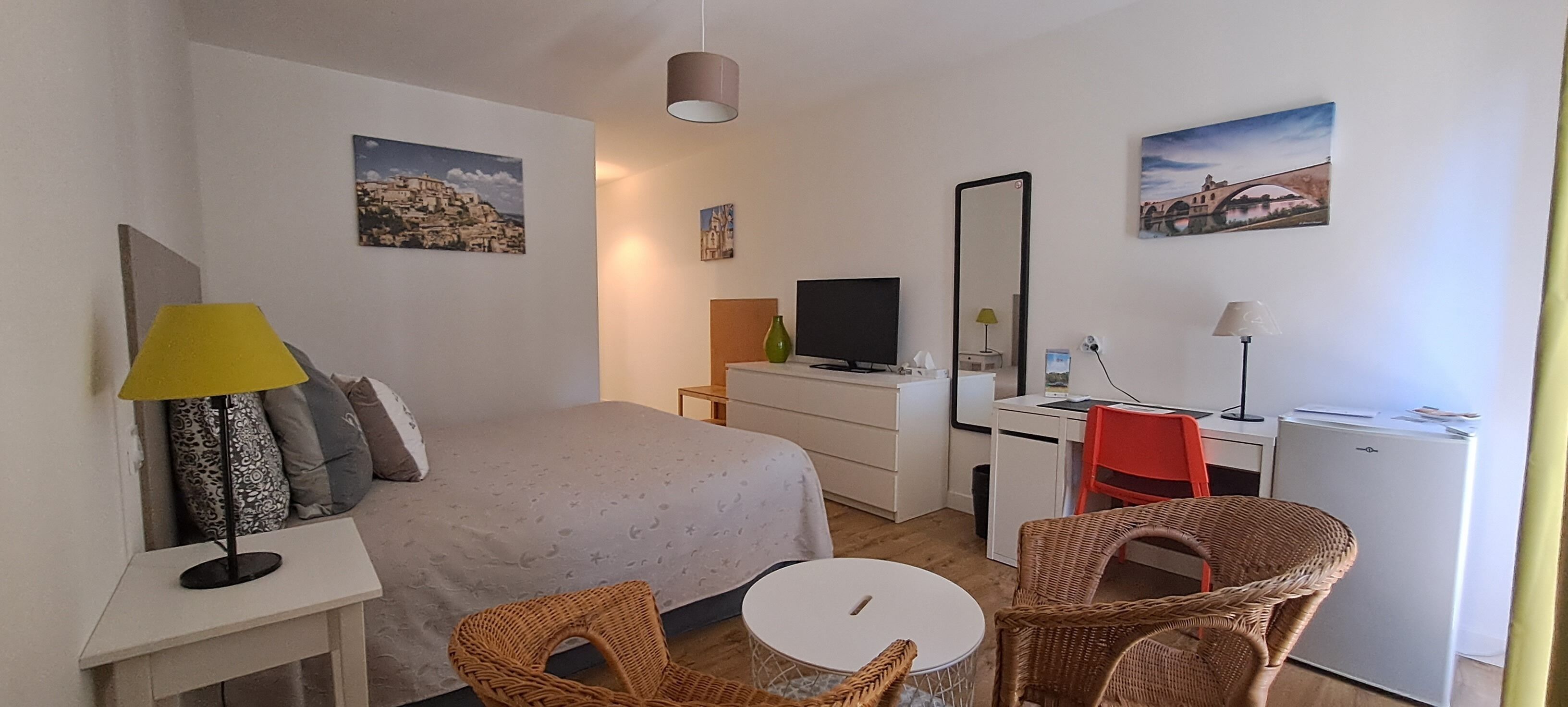 A hotel room with a bed, a desk, a TV, a mirror, and wicker chairs.