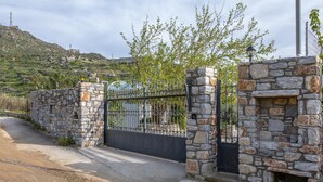 A gated entrance with stone walls and a metal gate, leading to a property with a stone fireplace and a view of a hilly landscape. - Villa Parisi Naxos Deluxe 1 (Agkidia)