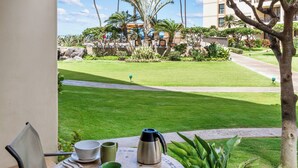 A table with a coffee pot, cups, and a bowl on a balcony overlooking a well-maintained lawn and a building in the background. - Sugar Beach 102 Gorgeous Remodeled Home Beachfront Condo Home (Kihei)