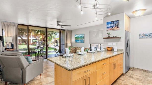 A modern kitchen with a granite countertop, wooden cabinets, and a large island. There is a dining area with a gray sofa and a table, and a view of a garden through large glass doors. - Sugar Beach 102 Gorgeous Remodeled Home Beachfront Condo Home (Kihei)