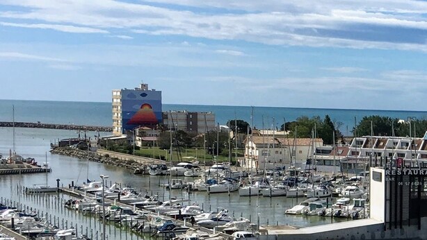 A marina with numerous boats docked, a prominent building with a colorful mural, and a restaurant sign. - Studio cabin 4 beds, sea view: beach 5 min + parking + air conditioning (Mauguio)