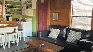 Una sala de estar acogedora con un sofá de cuero, una mesa de centro de madera y una cocina con armarios blancos y una puerta roja.