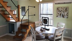 Una escalera de madera que lleva al segundo piso, un comedor con una mesa redonda y sillas, y un ventilador de techo.