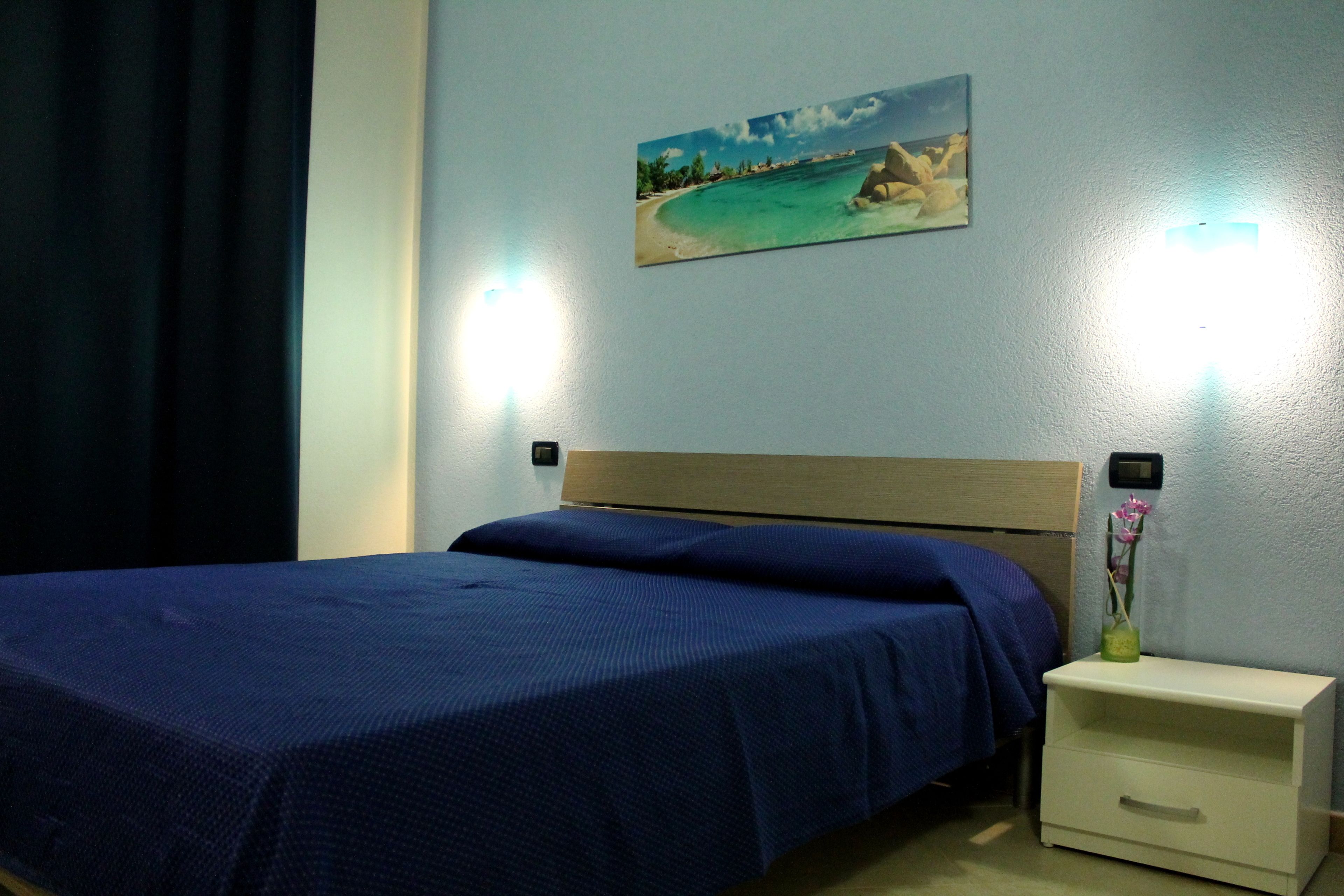 A neatly made bed with a blue bedspread in a room with a light blue wall, a painting of a beach, and a nightstand with a vase of flowers.