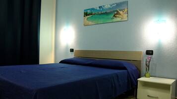 A neatly made bed with a blue bedspread in a room with a light blue wall, a painting of a beach, and a nightstand with a vase of flowers.