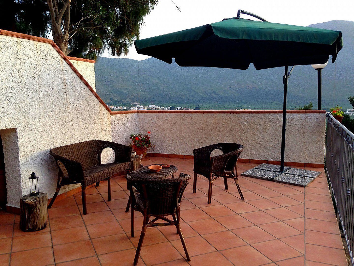 A terrace with wicker furniture, a green umbrella, and a view of distant mountains.
